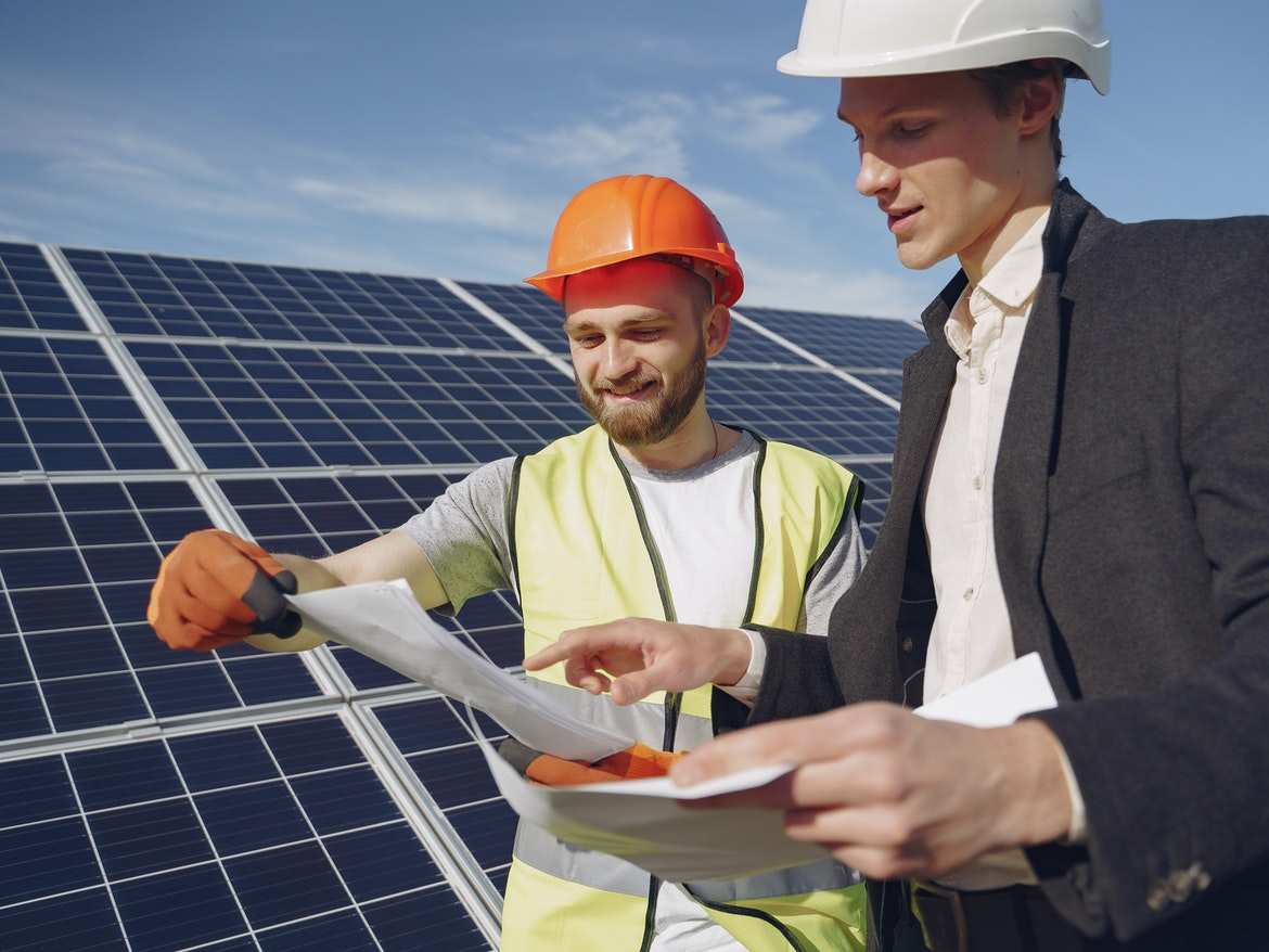 Két szakember védősisakban terveket egyeztet egy nagy napelemsor előtt — helyszíni energetikai átvilágítás