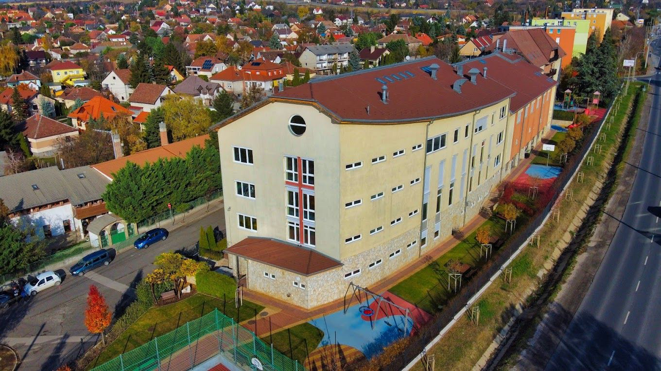 Iskolaépület légi fotója: többszintes intézményi épület játszóudvarral, sportpályával és fasorral, lakóövezeti környezetben
